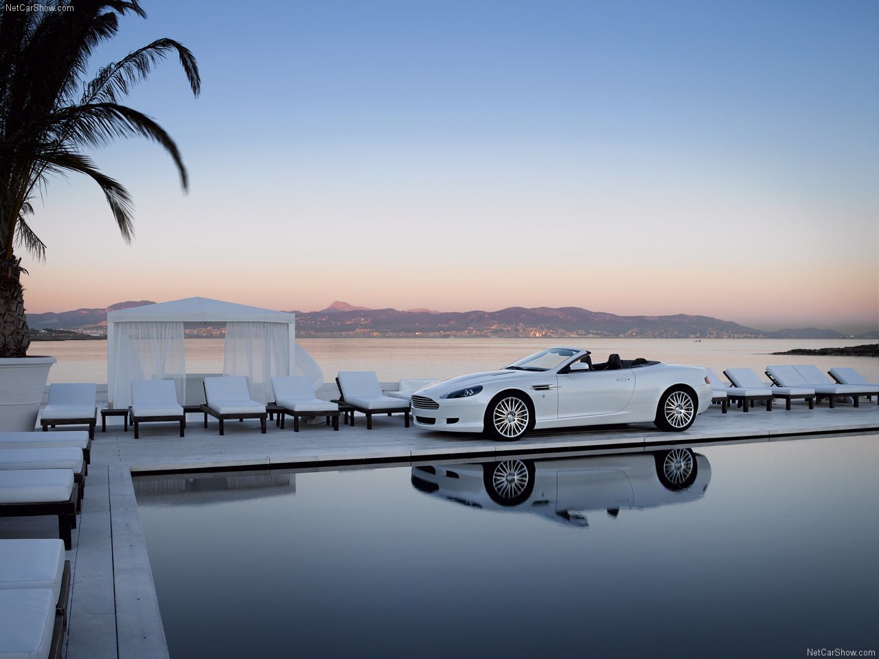 White Aston Martin vehicle convertible parked beside a tranquil pool with loungers at sunset, a palm tree and distant mountains reflected in the water.