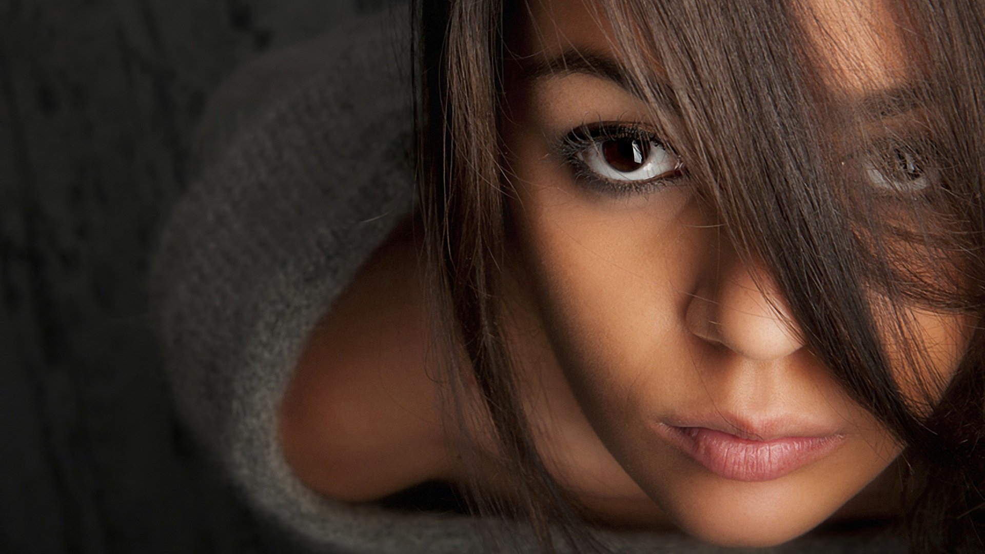 Close-up of a woman's face with hair partially covering one eye, showcasing expressive eyes and smooth skin against a dark, blurred background.