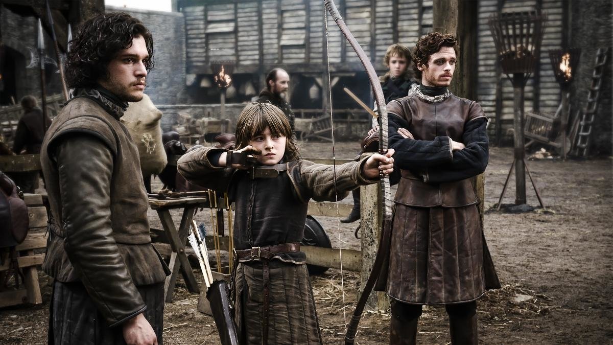 Three characters from the TV show Game of Thrones are in a medieval setting. A young boy draws a bow, while two men observe him, all dressed in period attire.