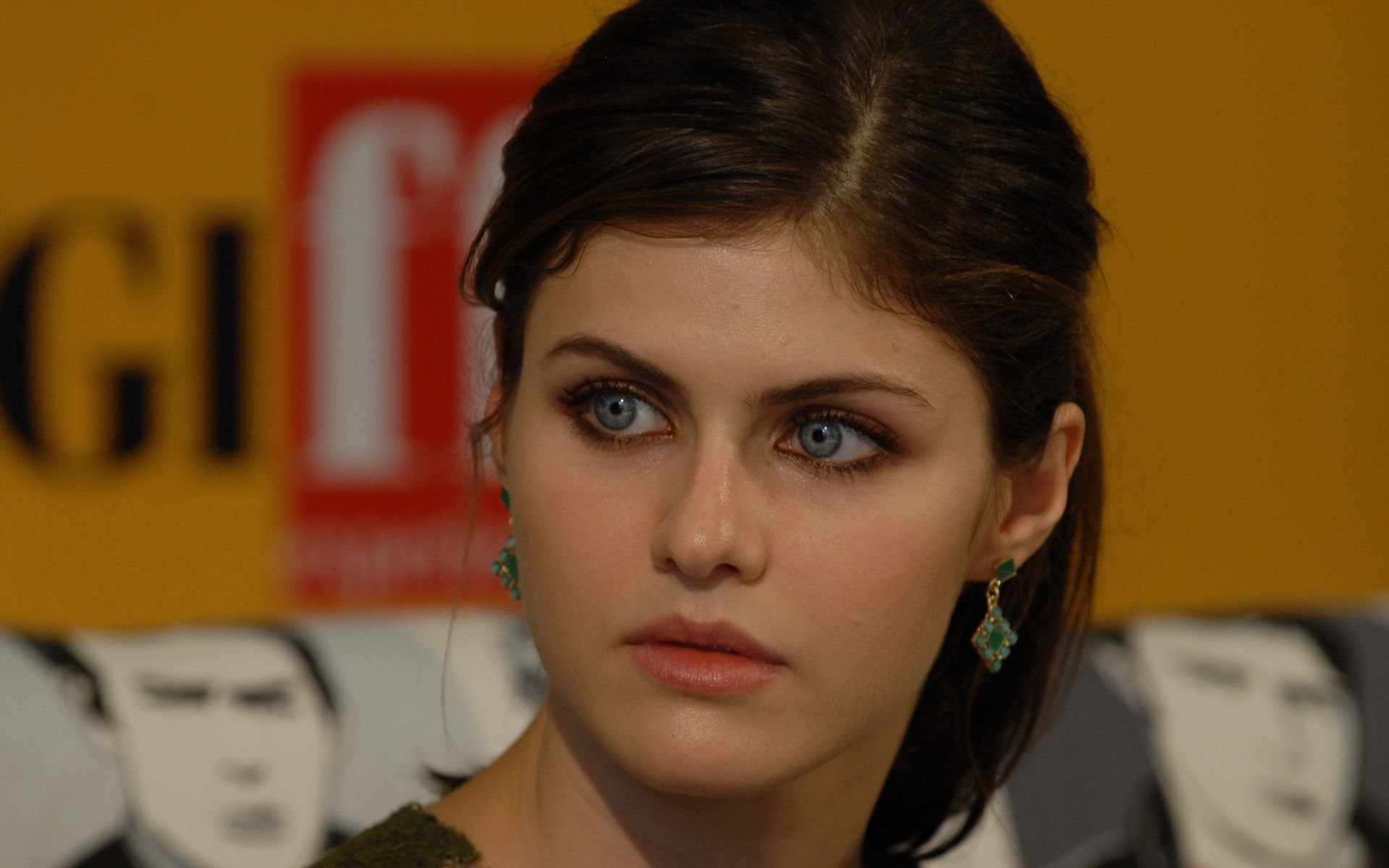 Portrait of celebrity Alexandra Daddario with striking blue eyes, dark hair, and green earrings, set against a blurred background featuring bold text.