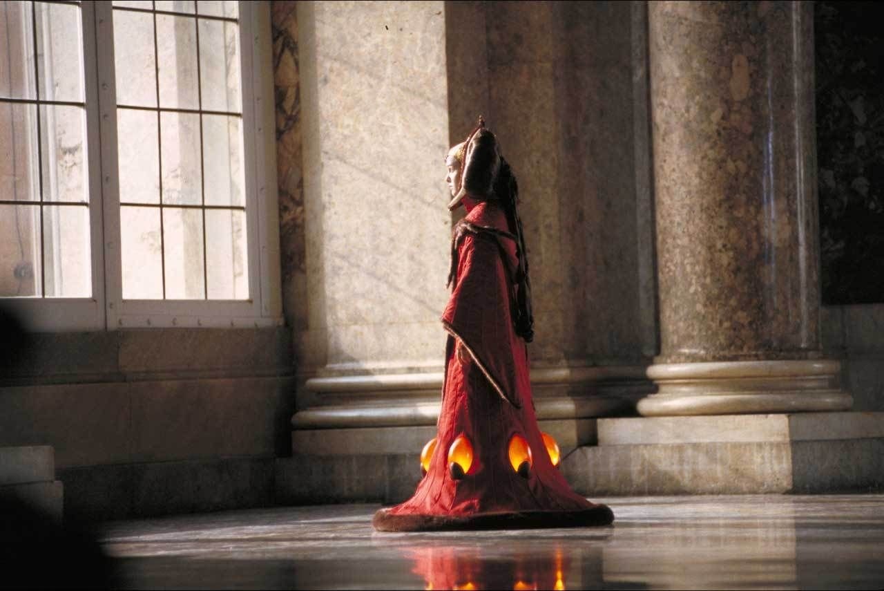 A cloaked figure stands in a grand space, illuminated by soft light, evoking the dramatic scene from *Star Wars Episode I: The Phantom Menace*.