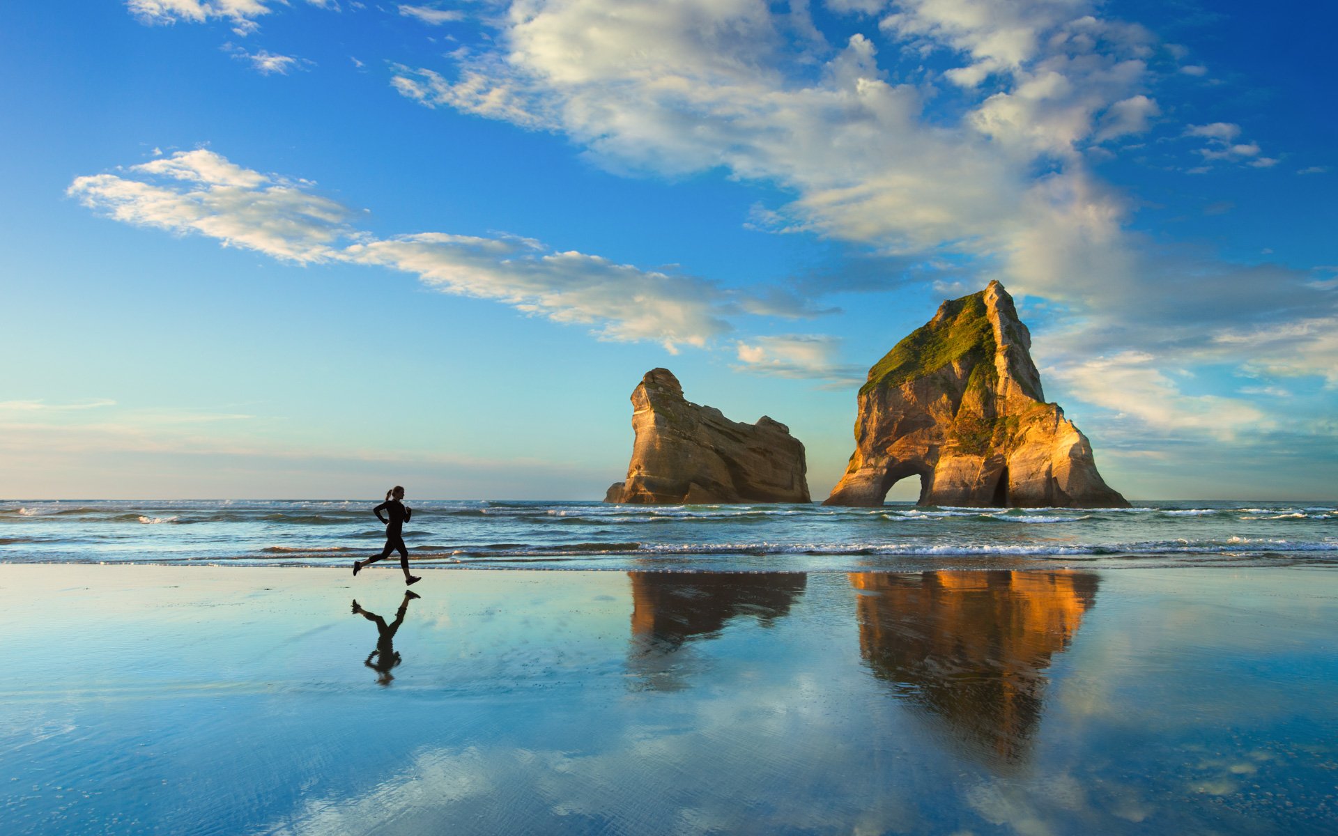 Photography captures a person running along a reflective beach shoreline with large rock formations under a blue sky scattered with clouds.