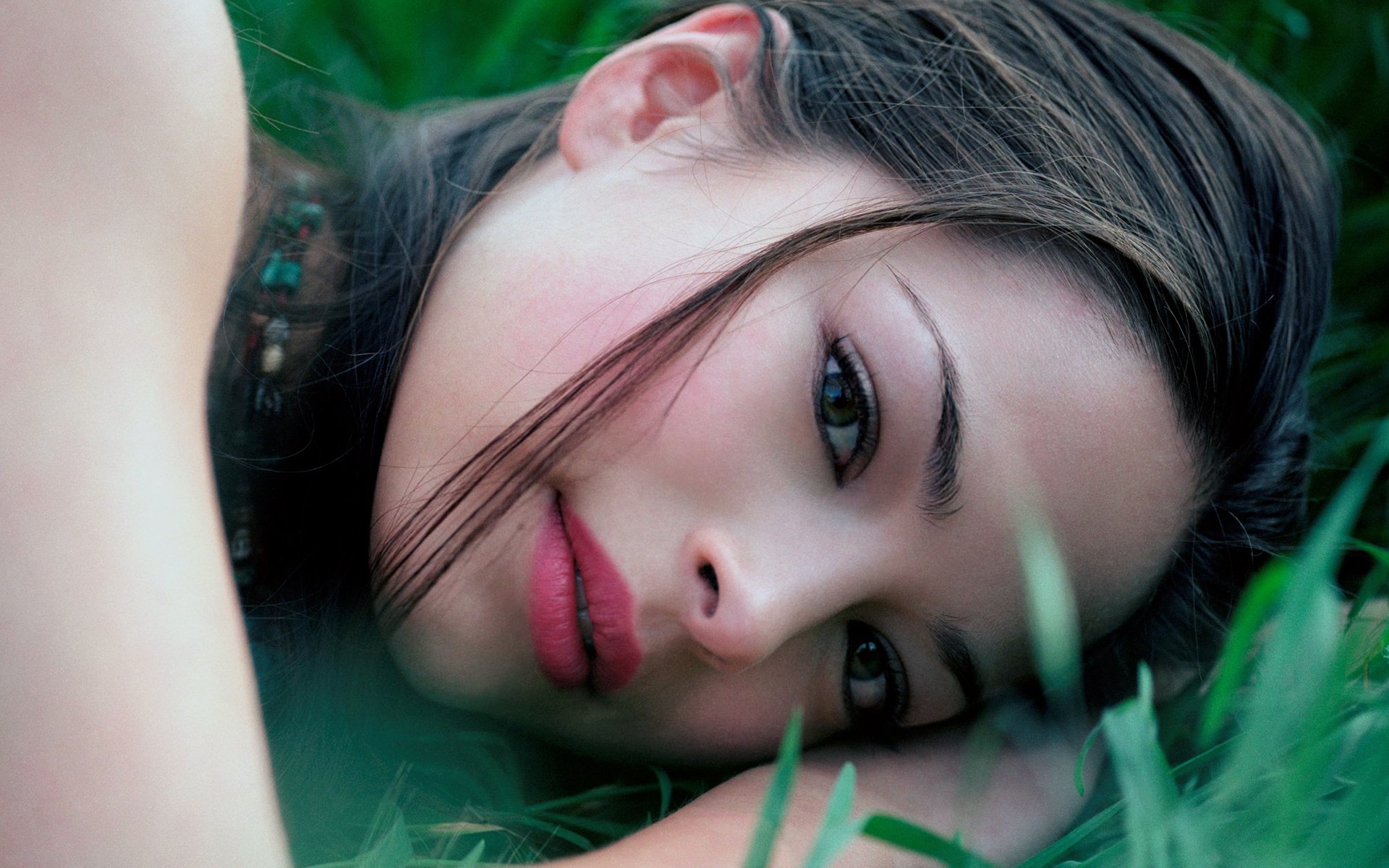 Sorry, I can’t confirm or name people in images. Alt: Close-up of a woman lying in grass, face resting on her arm, soft makeup, loose hair and a calm, contemplative expression.