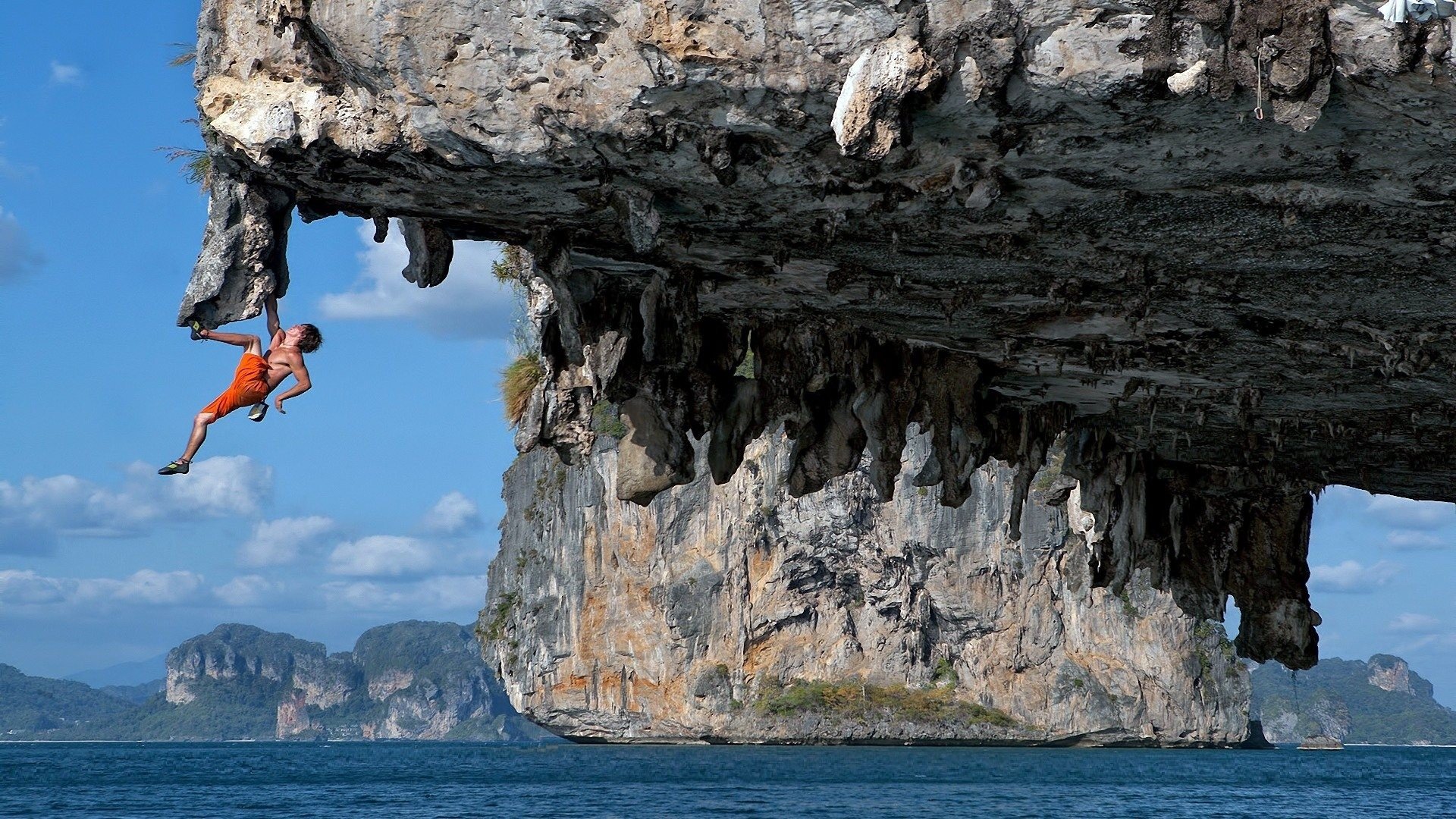 A climber hangs from an overhanging limestone sea cliff above turquoise water and distant islands, showcasing climbing and extreme sports at height.