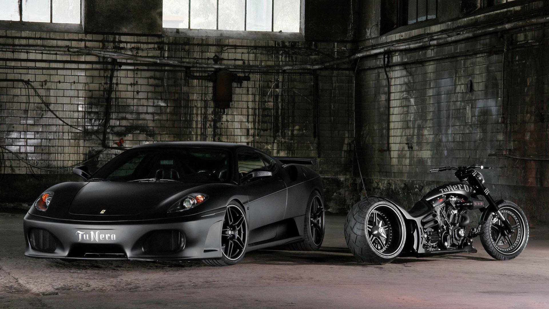 A sleek black Ferrari sports car and a custom black motorcycle parked side by side inside a dimly lit industrial garage.