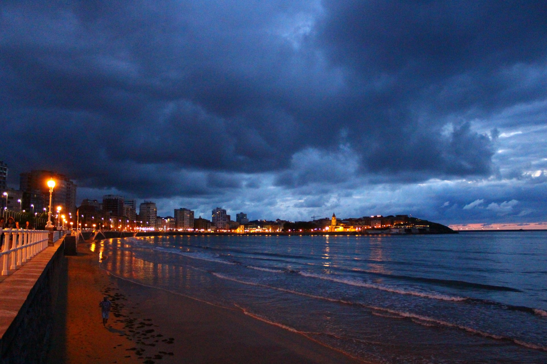 Twilight beach landscape with a man-made promenade and city skyline along the shore, glowing lights reflecting on the calm sea beneath brooding clouds.
