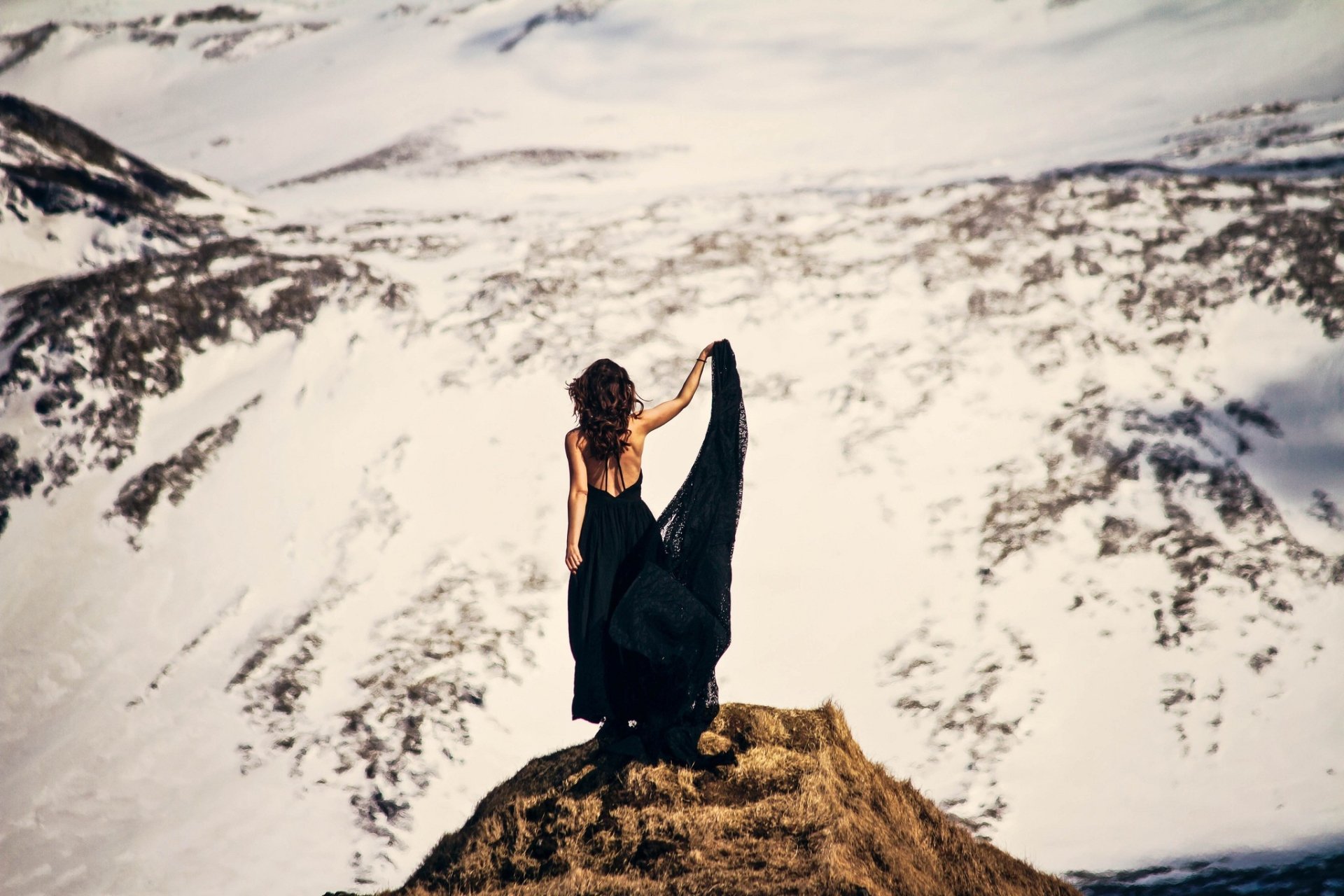 Brunette woman model stands on a mountain peak, viewed from the rear, holding a flowing black fabric against a snowy mountainous backdrop.