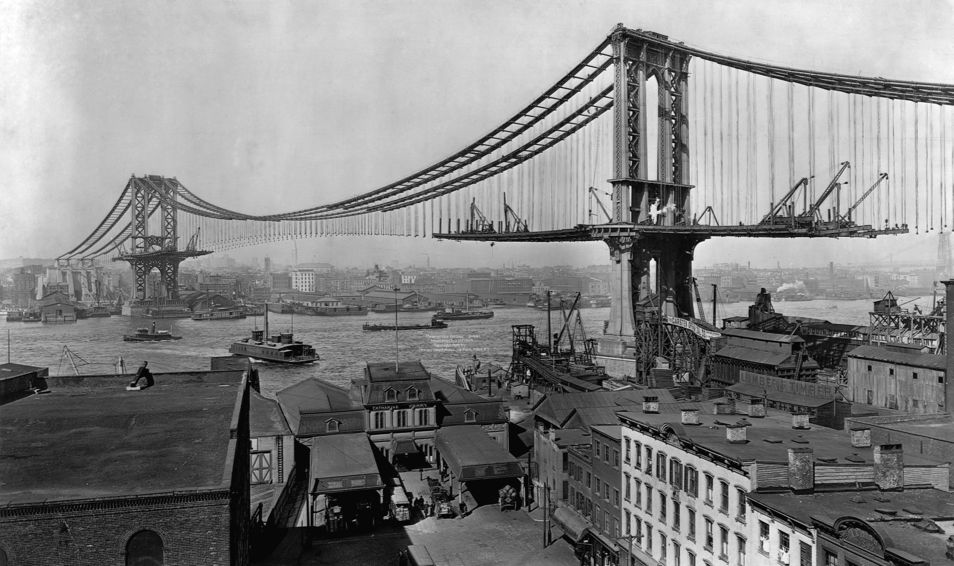 Black & white photography capturing the early construction phase of a large suspension bridge over a busy river with boats and industrial buildings in the foreground.