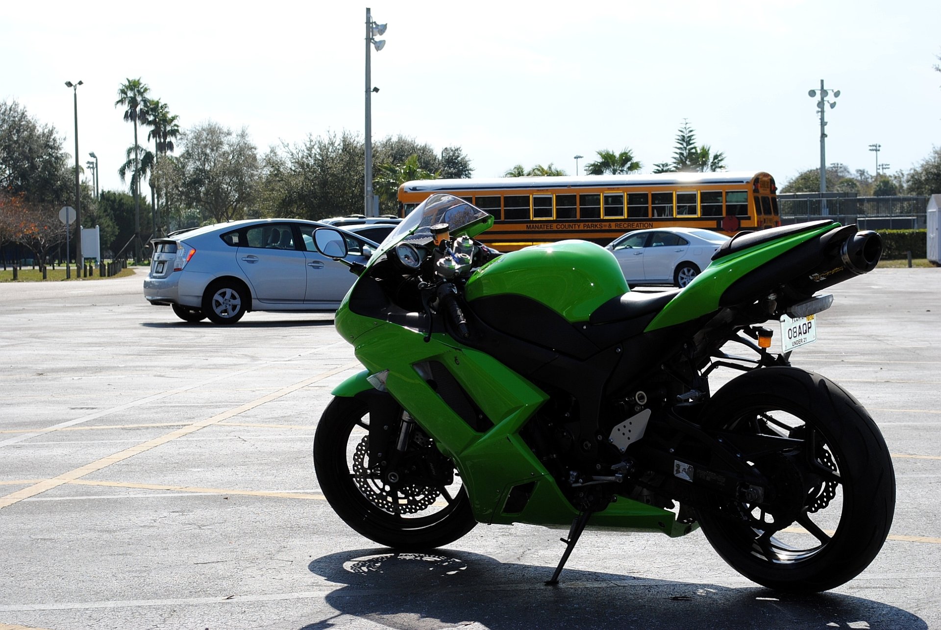 A vibrant green Kawasaki motorcycle stands in a parking lot, with a silver car and a school bus visible in the background under clear skies.