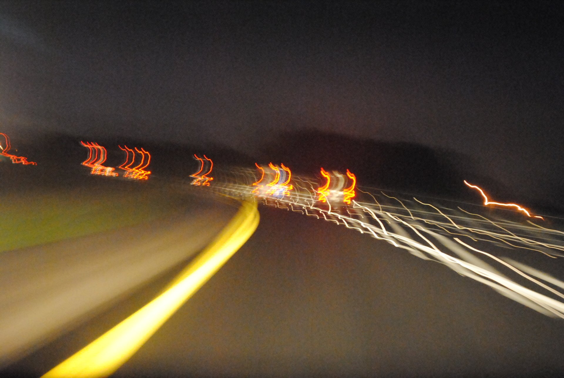 A dynamic night scene featuring a blurred highway, showcasing light trails from vehicles in motion. The photograph emphasizes creative manipulation of light and movement.