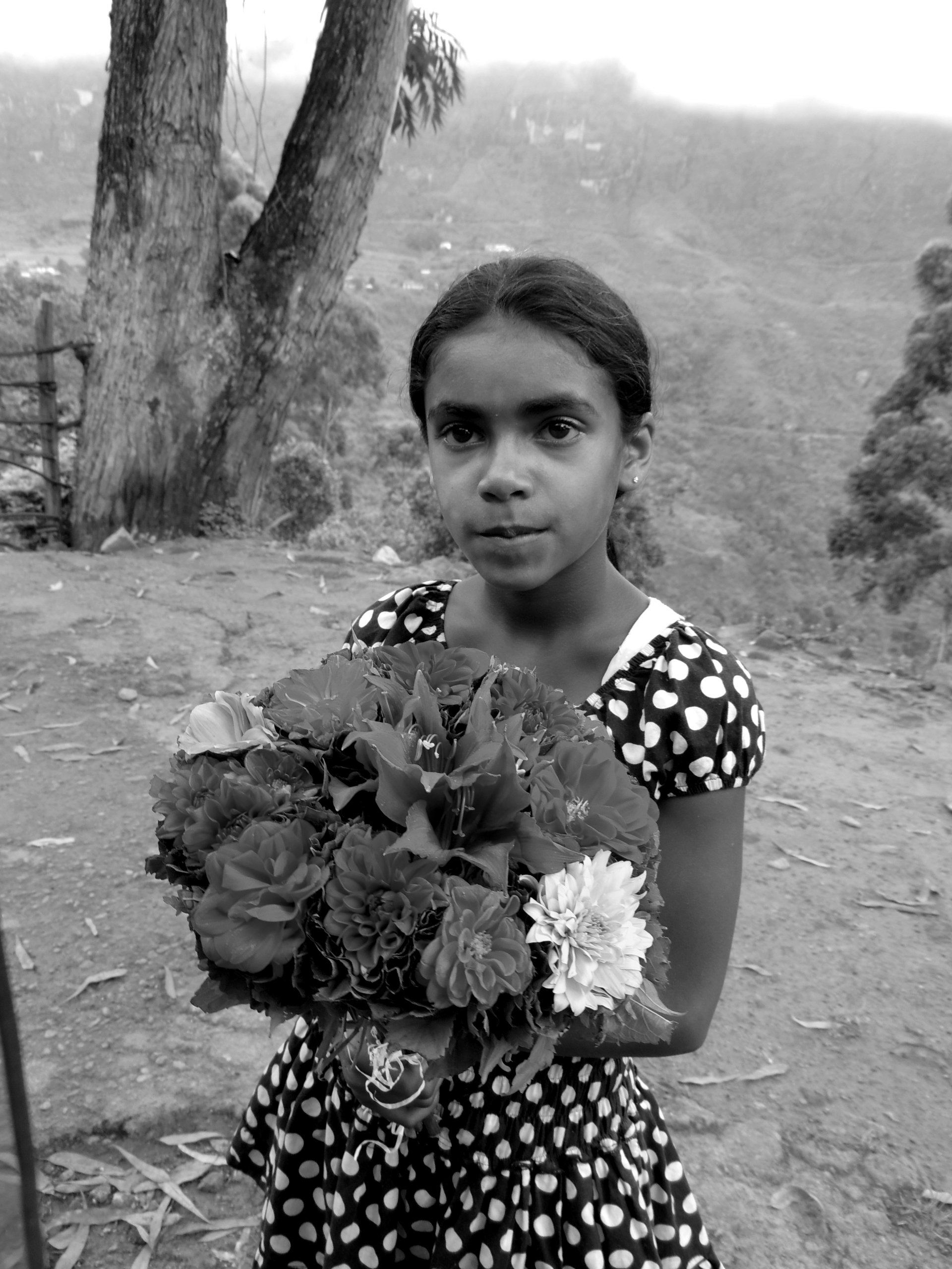 Black and white photography of a young girl holding a large bouquet of flowers outdoors with trees and hills in the background.