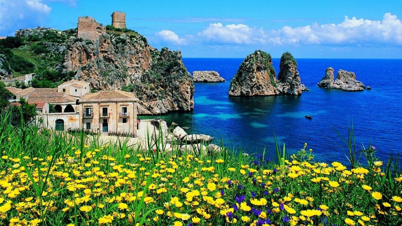 A vibrant flower-filled foreground leads to historic houses overlooking the rocky coast and deep blue ocean of Sicily, Italy.
