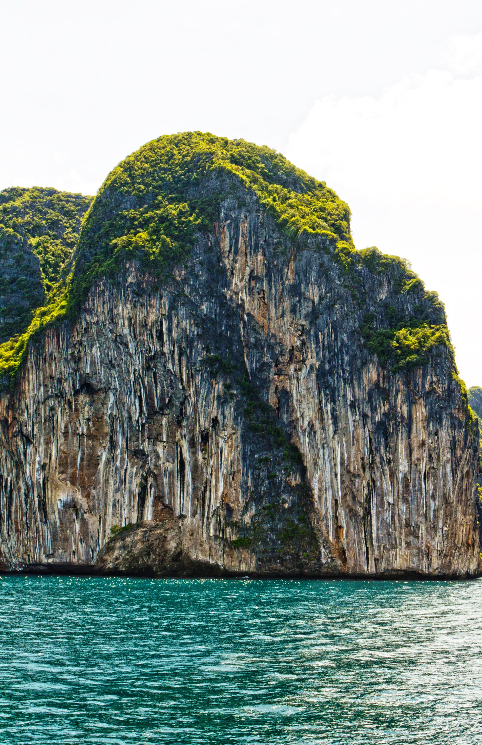 A towering cliff covered in lush greenery rises sharply from the vibrant turquoise water, showcasing the stunning beauty of nature.