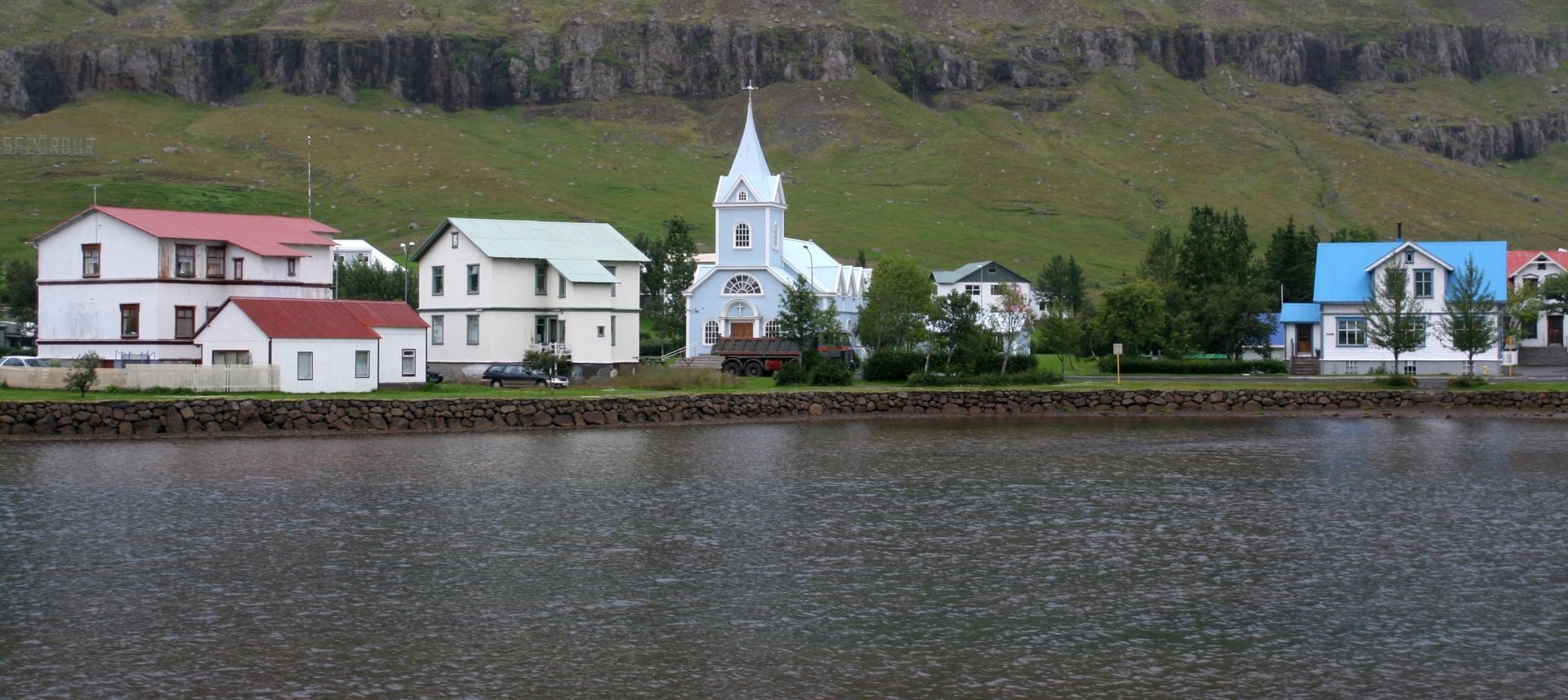 A man-made village with colorful houses and a white church along a waterfront, set against a backdrop of green hills and rocky cliffs.