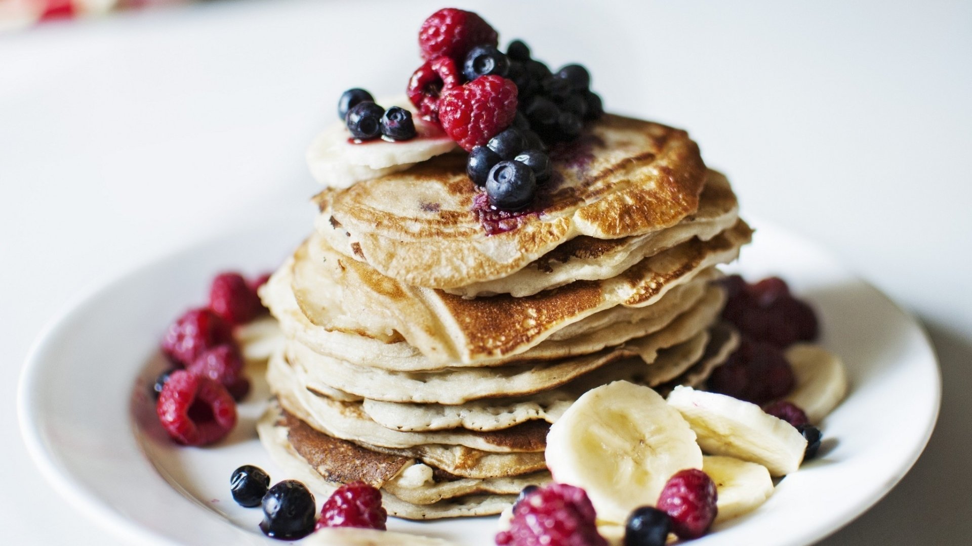 Stack of pancakes topped with raspberries, blueberries, and banana slices on a white plate.