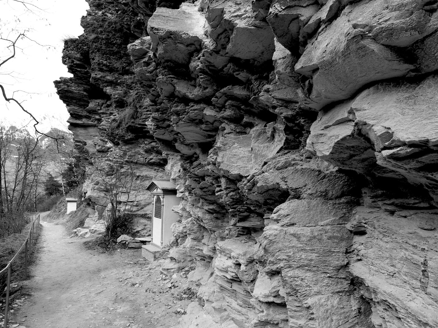 A rugged rock formation frames a winding path leading to small structures nestled at the base, showcasing the beauty of nature in a monochrome setting.