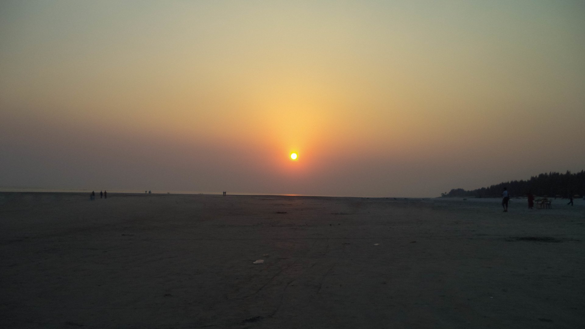 Photo of a beach at sunset with the sun low on the horizon, warm glow on calm water and a wide sandy shore dotted with distant silhouetted figures.