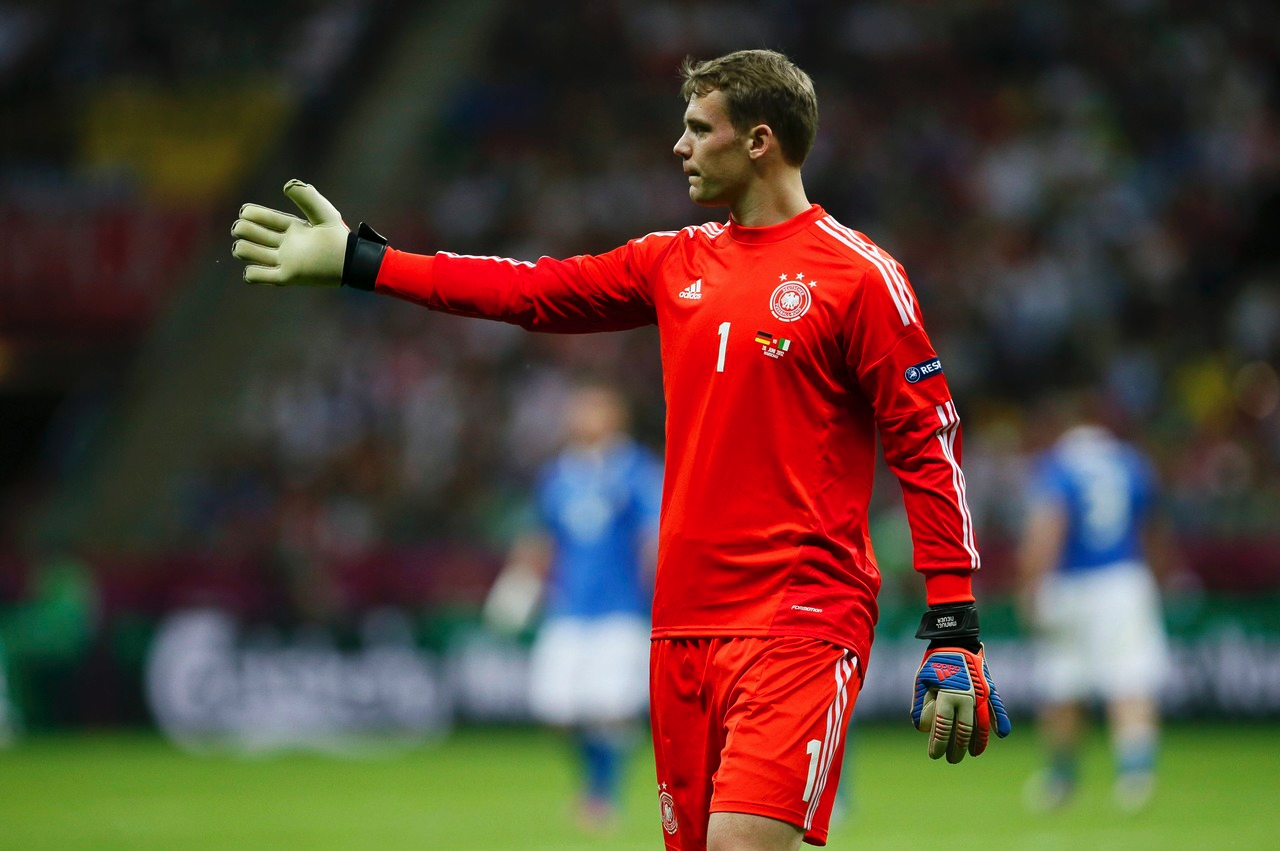 Manuel Neuer, the German soccer goalkeeper, directs play on the field while wearing his iconic red uniform during a match. The atmosphere is energetic with fans in the background.