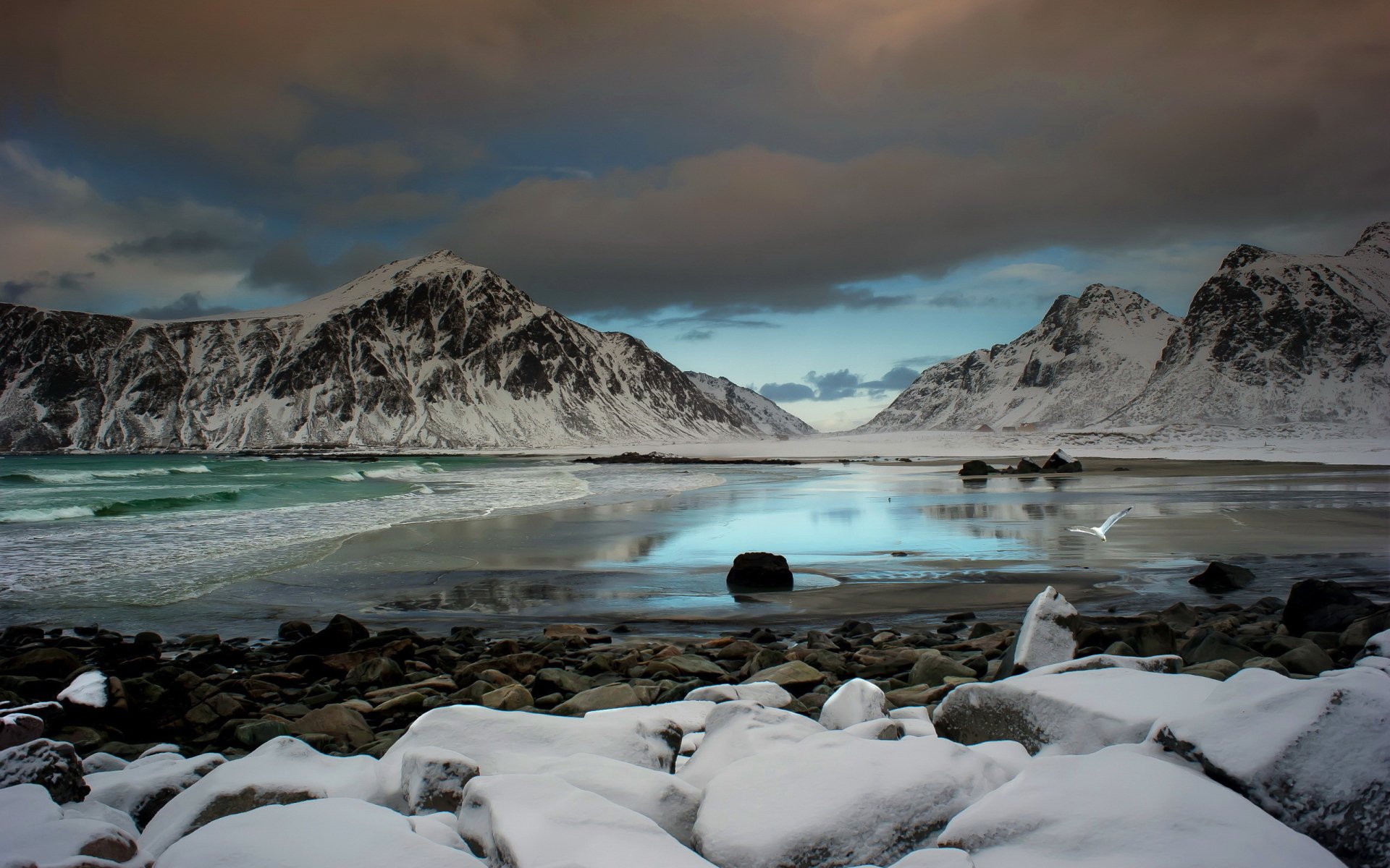 Snow-covered beach cove in a nature landscape, rugged mountains encircling turquoise tidal pools reflecting a dramatic cloudy sky.