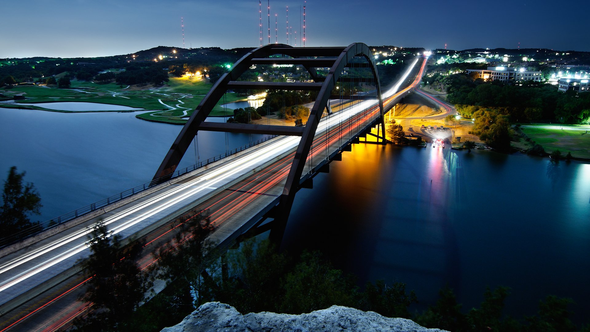 A time-lapse photograph captures the illuminated contours of a bridge spanning a river, with light trails from vehicles against a backdrop of a serene night skyline.