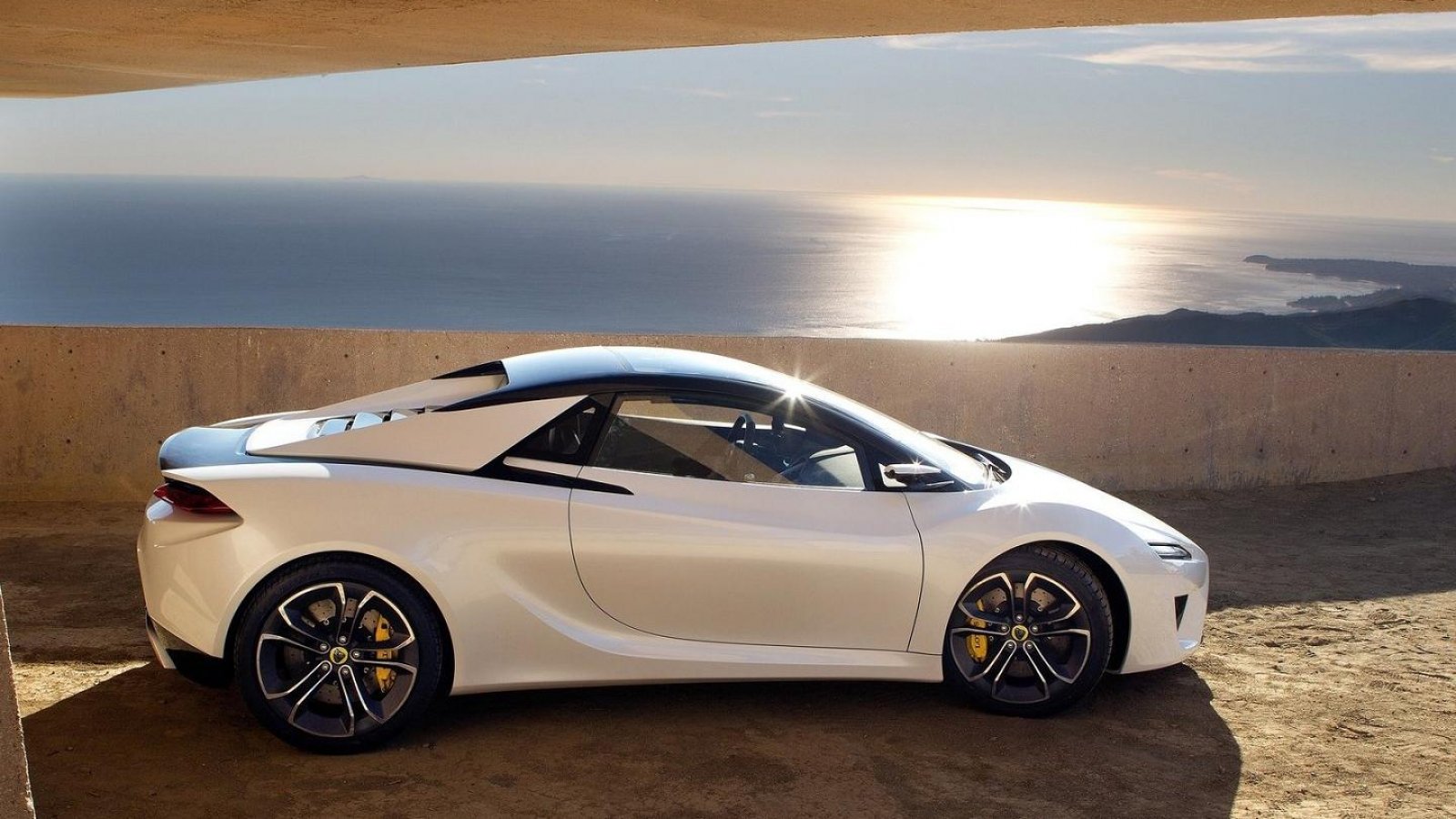 White Lotus sports vehicle parked on a coastal overlook at sunset, side profile revealing sleek aerodynamic lines and multi-spoke alloy wheels.