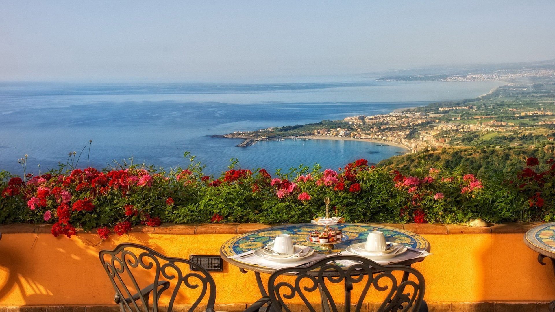 Photography captures a vibrant coastline view from a terrace with wrought-iron chairs, a table set for tea, and bright red flowers in the foreground.
