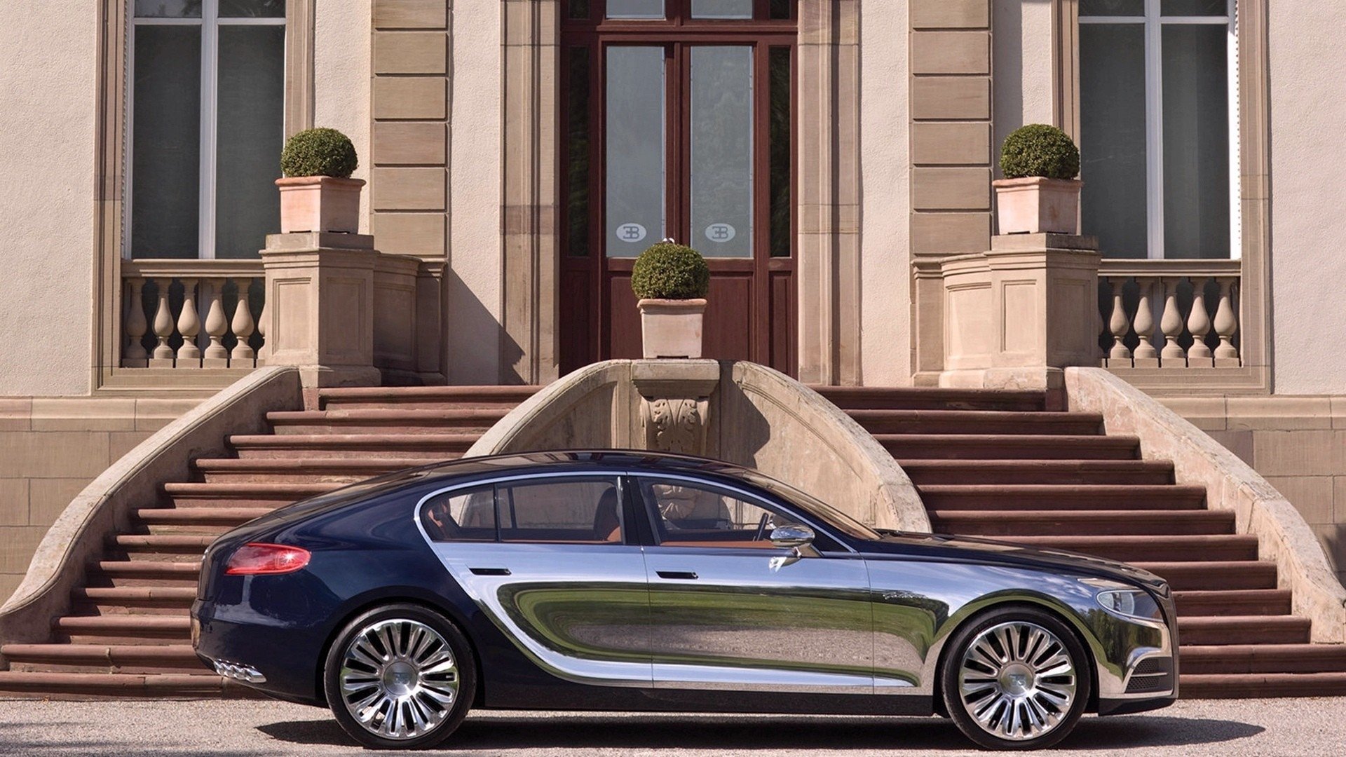 Side view of a Bugatti Galibier vehicle — a luxury car — parked before grand stone architecture with sweeping steps, balustrades and trimmed topiary.