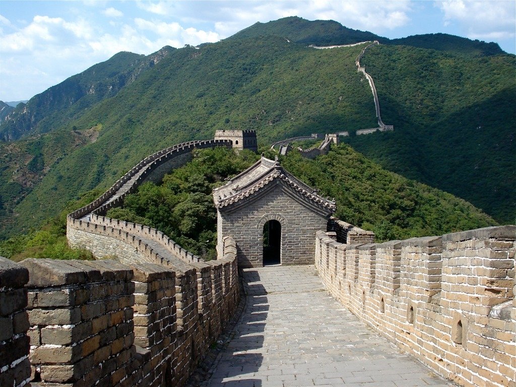 The Great Wall of China, a man-made structure, stretches across green mountainous terrain under a partly cloudy sky.