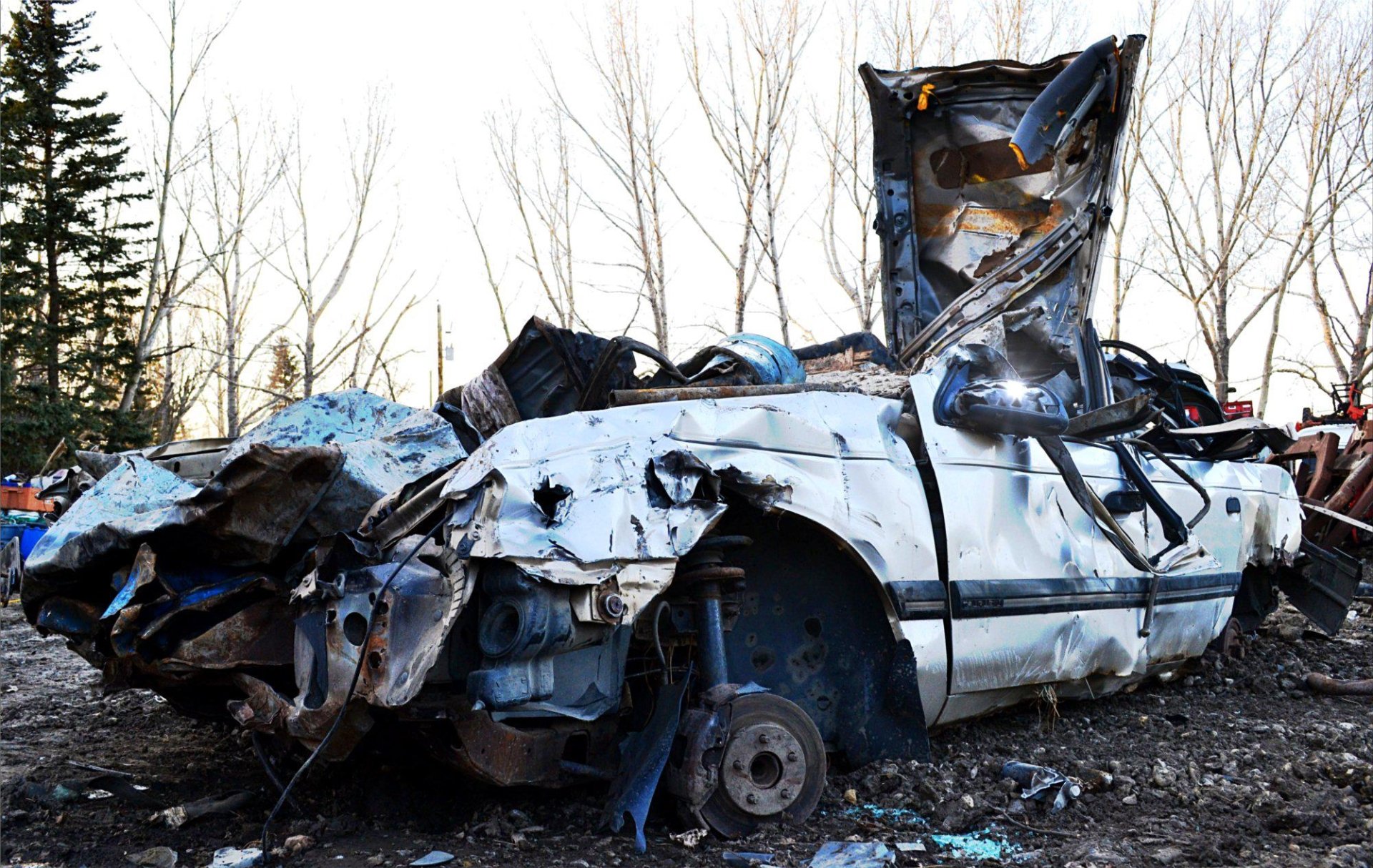 A severely wrecked white car with extensive damage is abandoned outdoors near leafless trees.