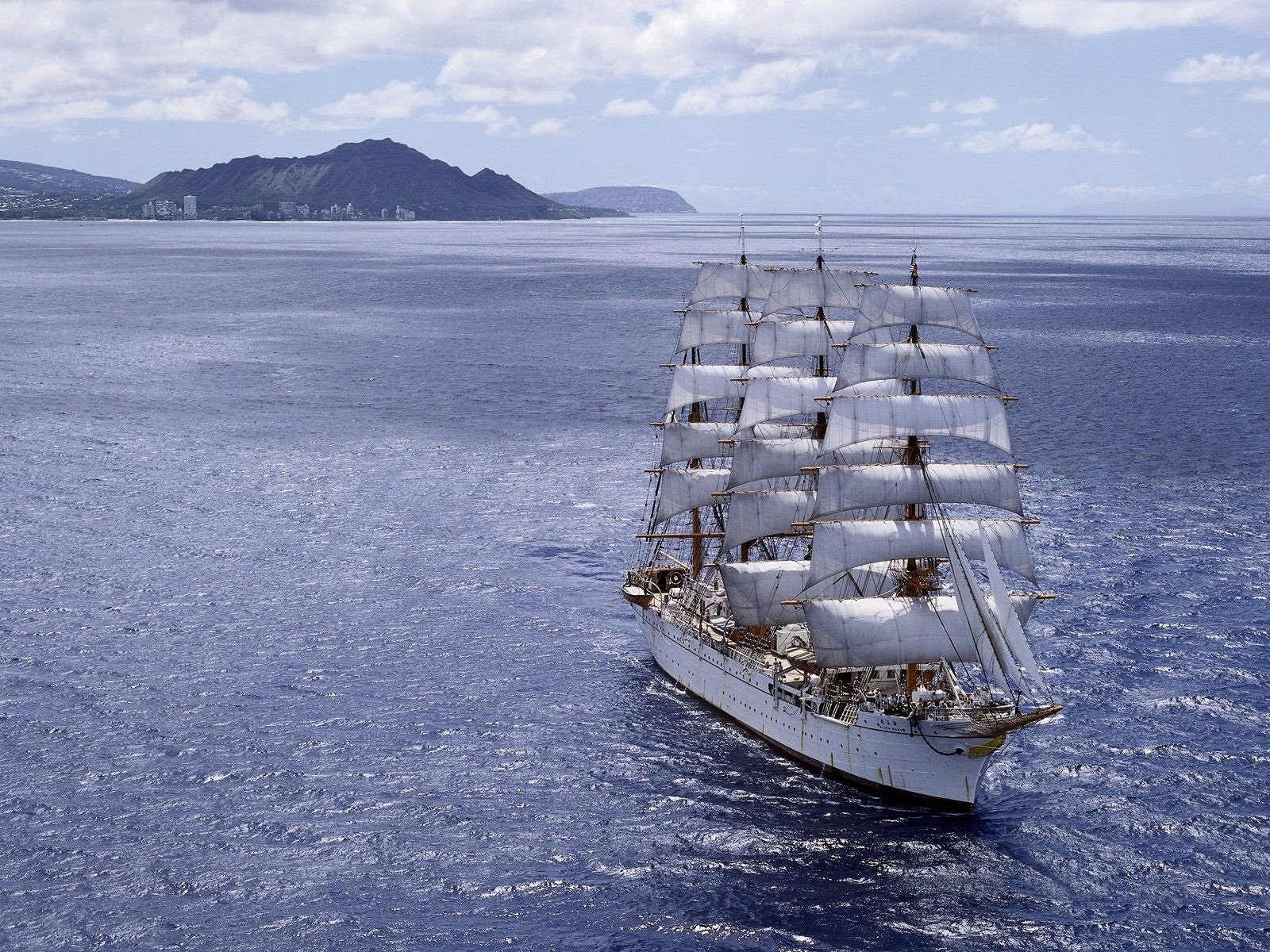 A majestic ship with multiple sails glides through the shimmering blue ocean, with a distant hilly coastline and cloudy sky in the background.