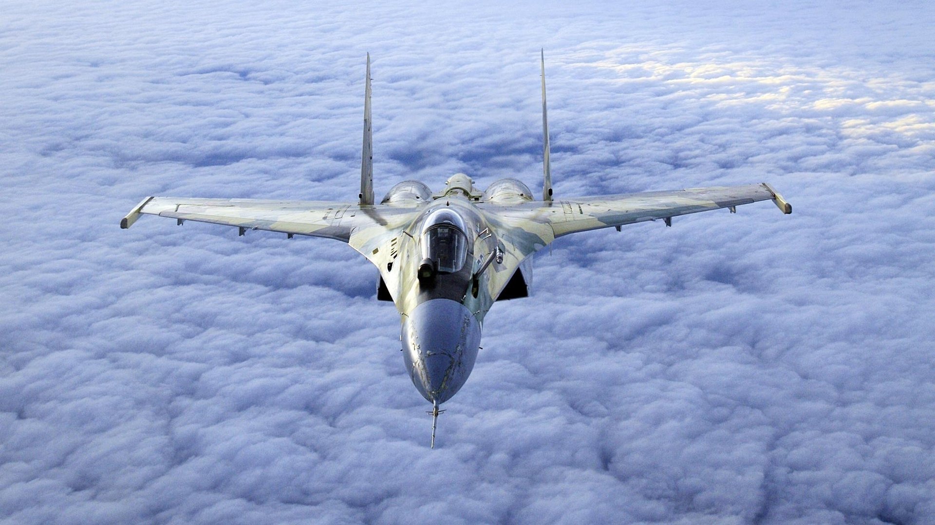A military Sukhoi Su-35 fighter jet flying above a layer of clouds, captured from the front with its wings spread wide.
