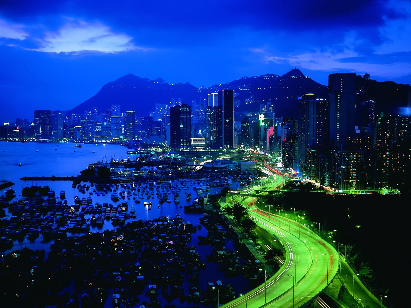 A stunning night view of Rio de Janeiro showcases a vibrant skyline, illuminated buildings, and winding roads alongside the waterfront, framed by mountains under a dramatic sky.
