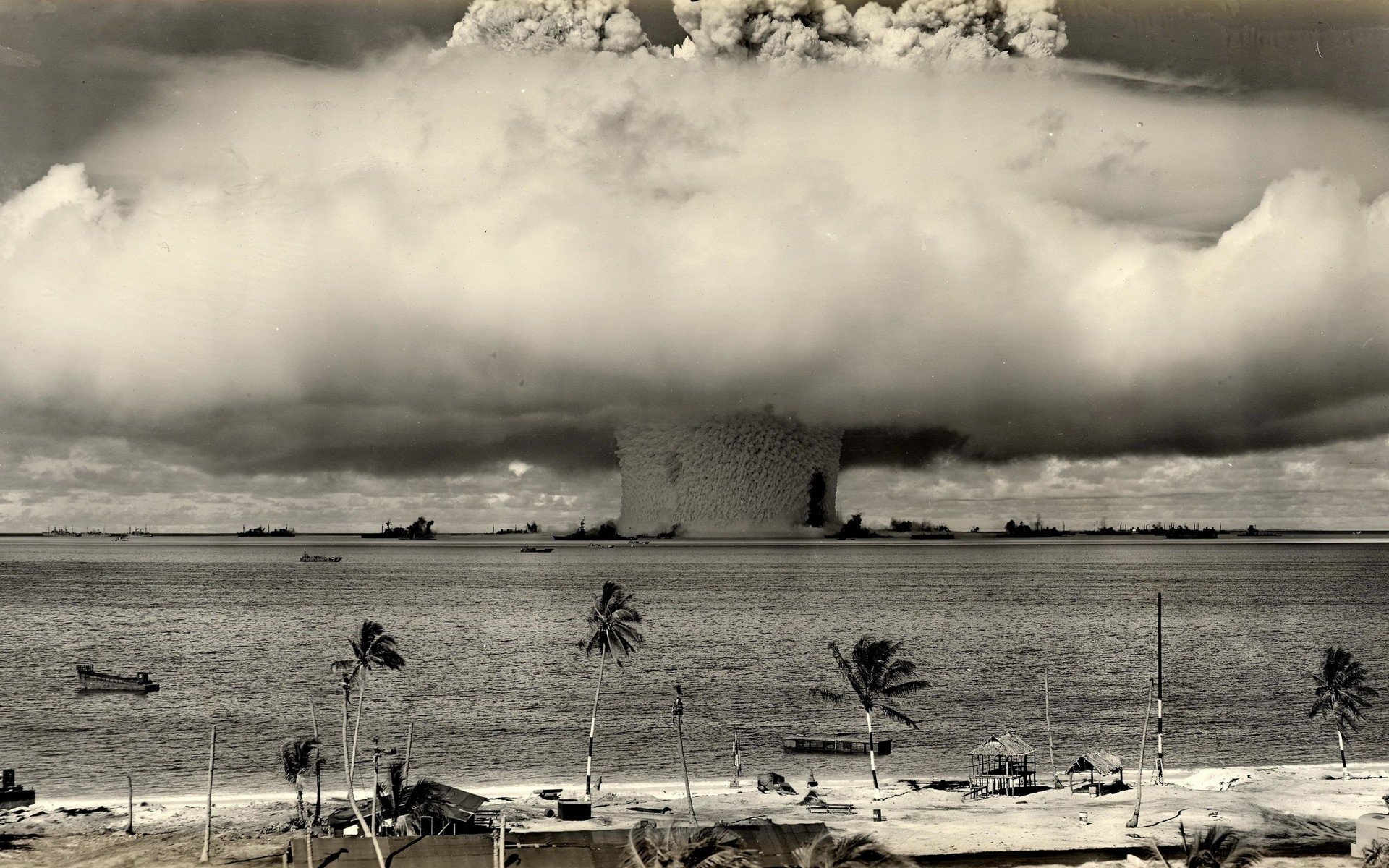 A massive military explosion creates a large mushroom cloud rising over the water near a coastal area with palm trees and buildings.