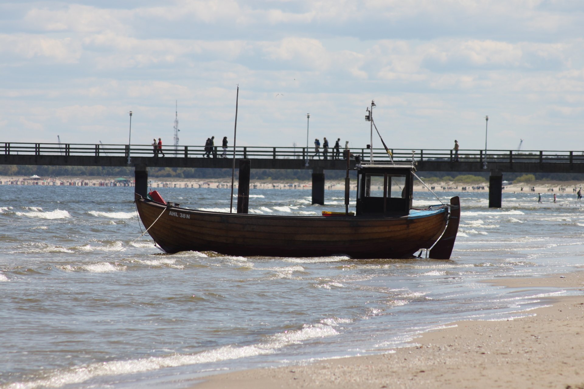 A wooden boat, a type of water vehicle, rests near the shore with a pier and people in the background under a partly cloudy sky.