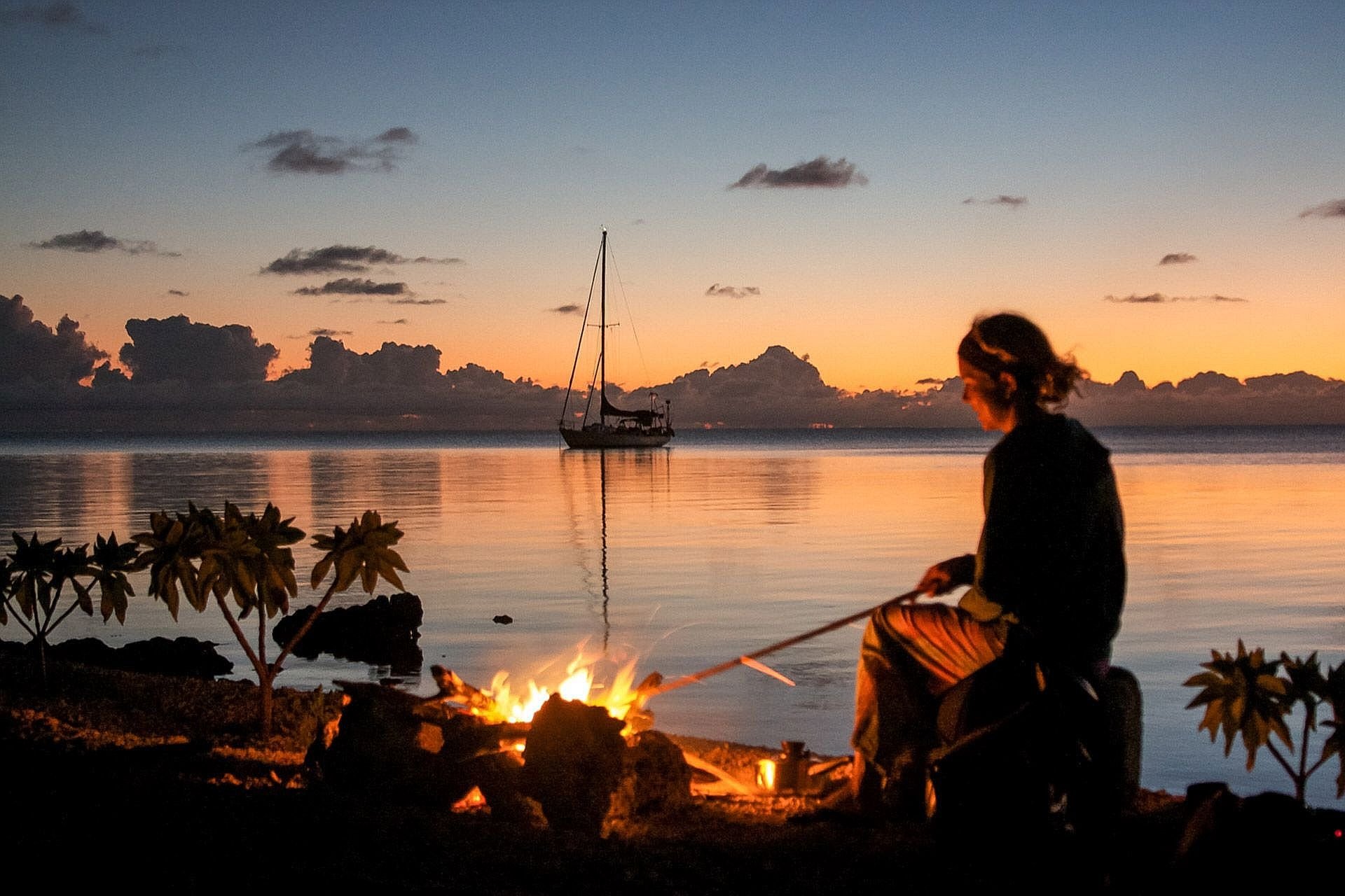 A woman named Lizzy Clark sits by a campfire on the beach at sunset, with a sailboat anchored in the calm waters behind her, surrounded by tropical plants.