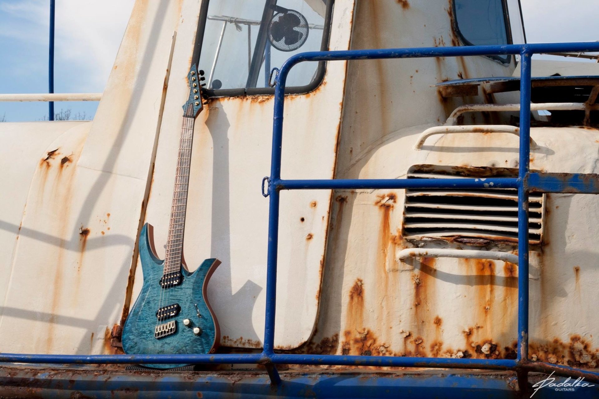 A blue electric guitar resting against the railing of a weathered, rust-streaked ship, blending elements of music and industrial texture.