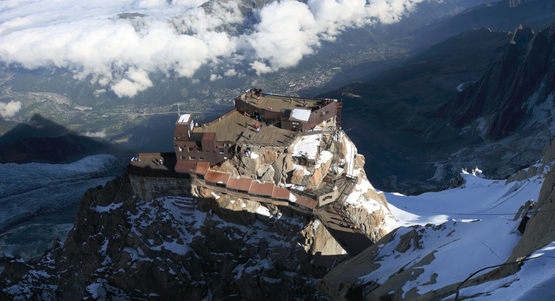 An aerial view of a majestic mountain landscape, featuring a weathered structure perched on a rocky ledge, surrounded by snow and clouds under a vast sky.