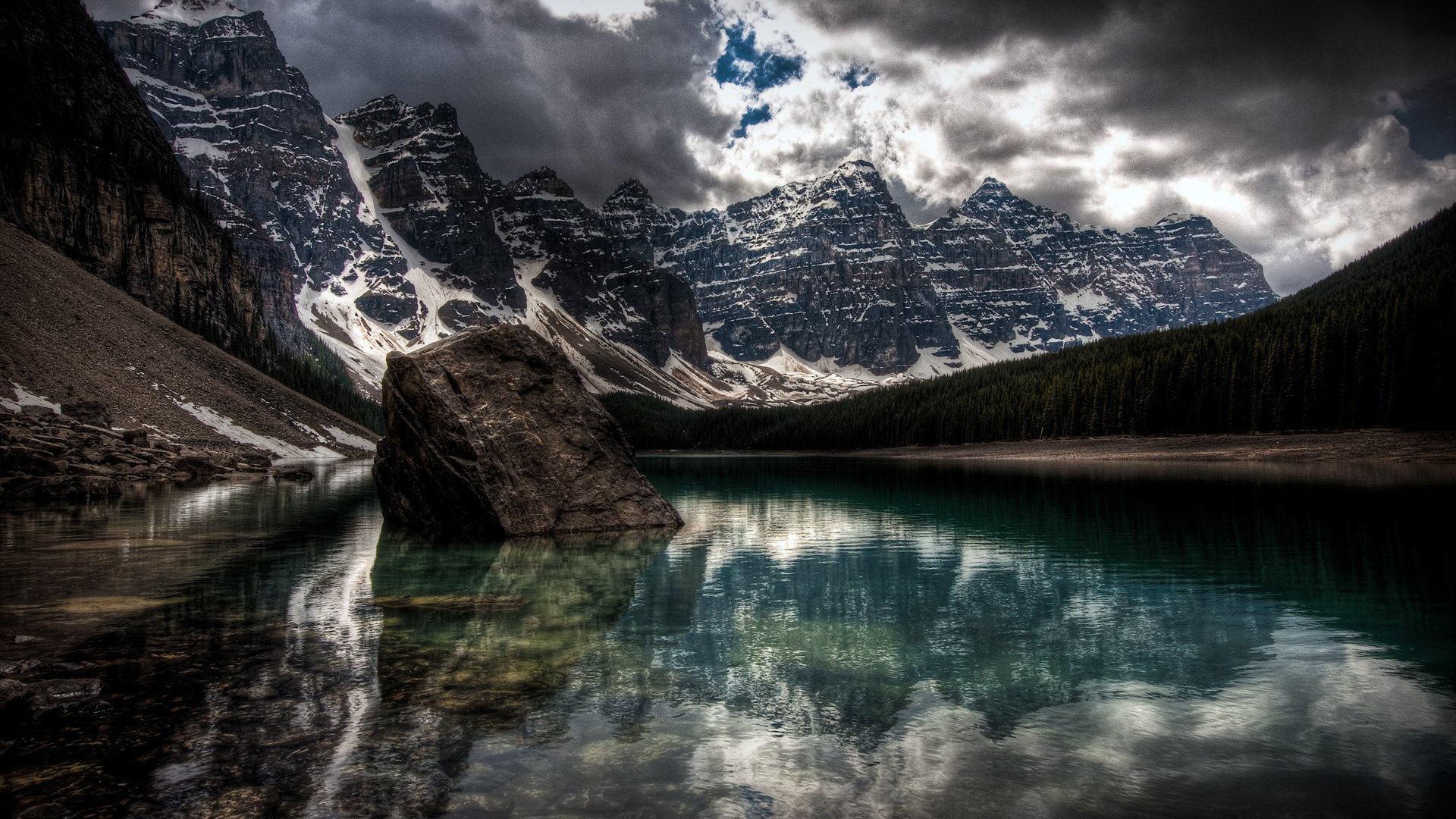 A stunning HDR photograph captures a serene lake reflecting dramatic mountains and a moody sky, surrounded by evergreen trees and rocky shores.