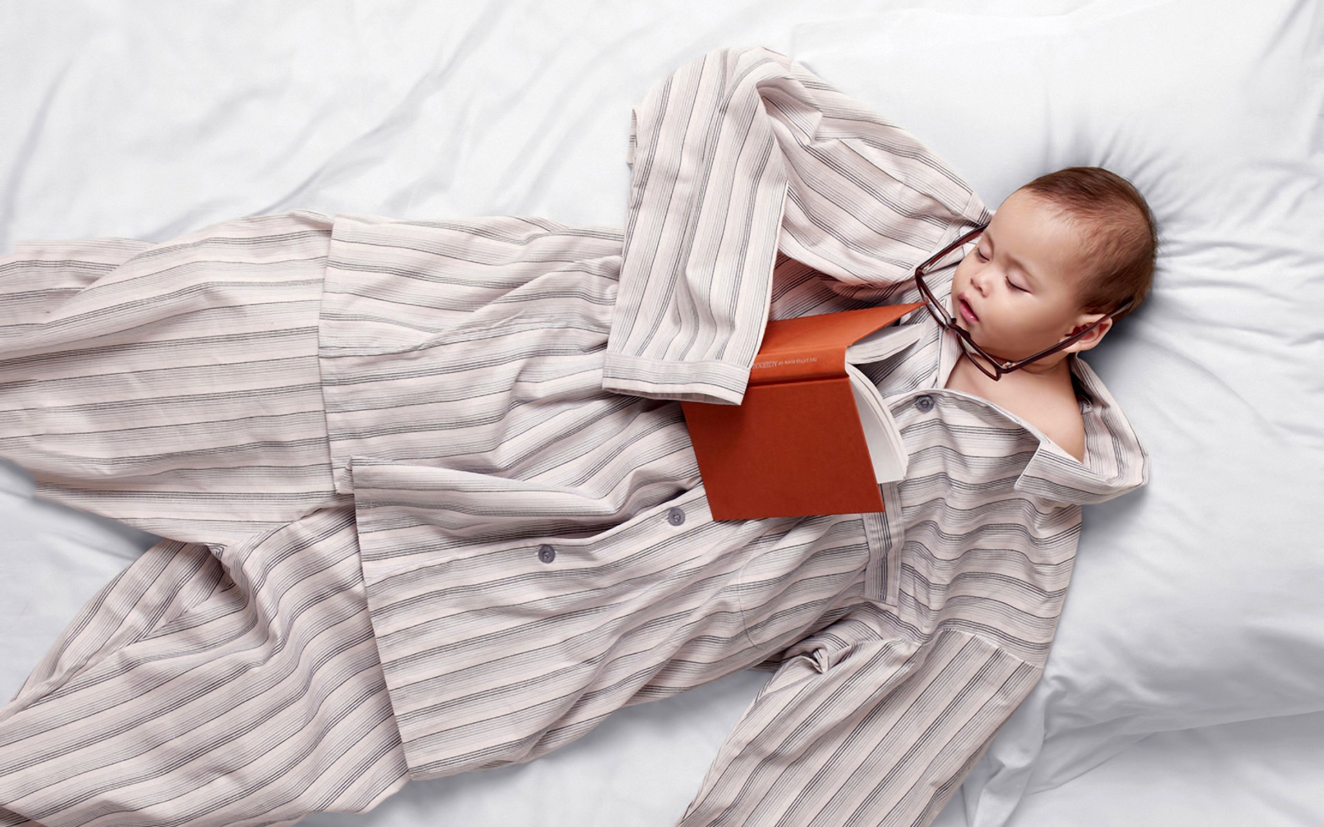A cute baby wearing glasses and striped pajamas lies on a bed, peacefully holding a book, embodying a funny and charming moment.