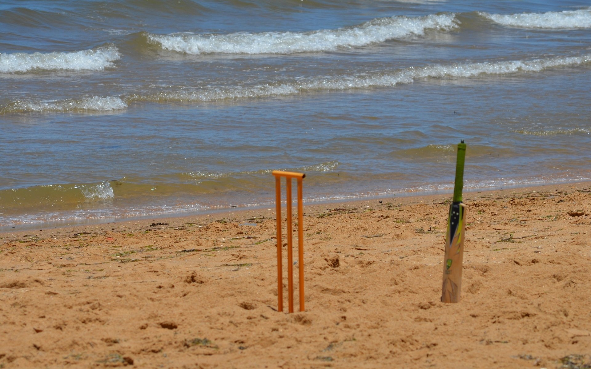 Two cricket stumps stand on a sandy beach, with gentle waves lapping at the shore. The scene captures a blend of sports and nature by the sea.