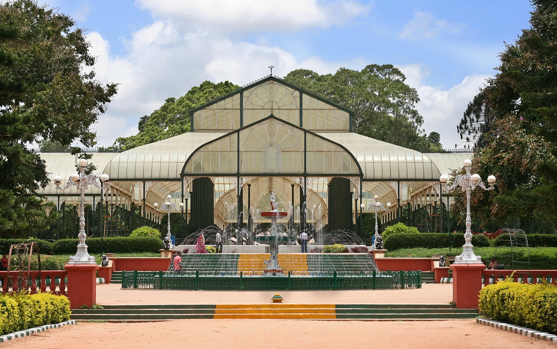 A man-made glass greenhouse stands at the center of a well-maintained garden with colorful flower beds and lamp posts lining the pathway.