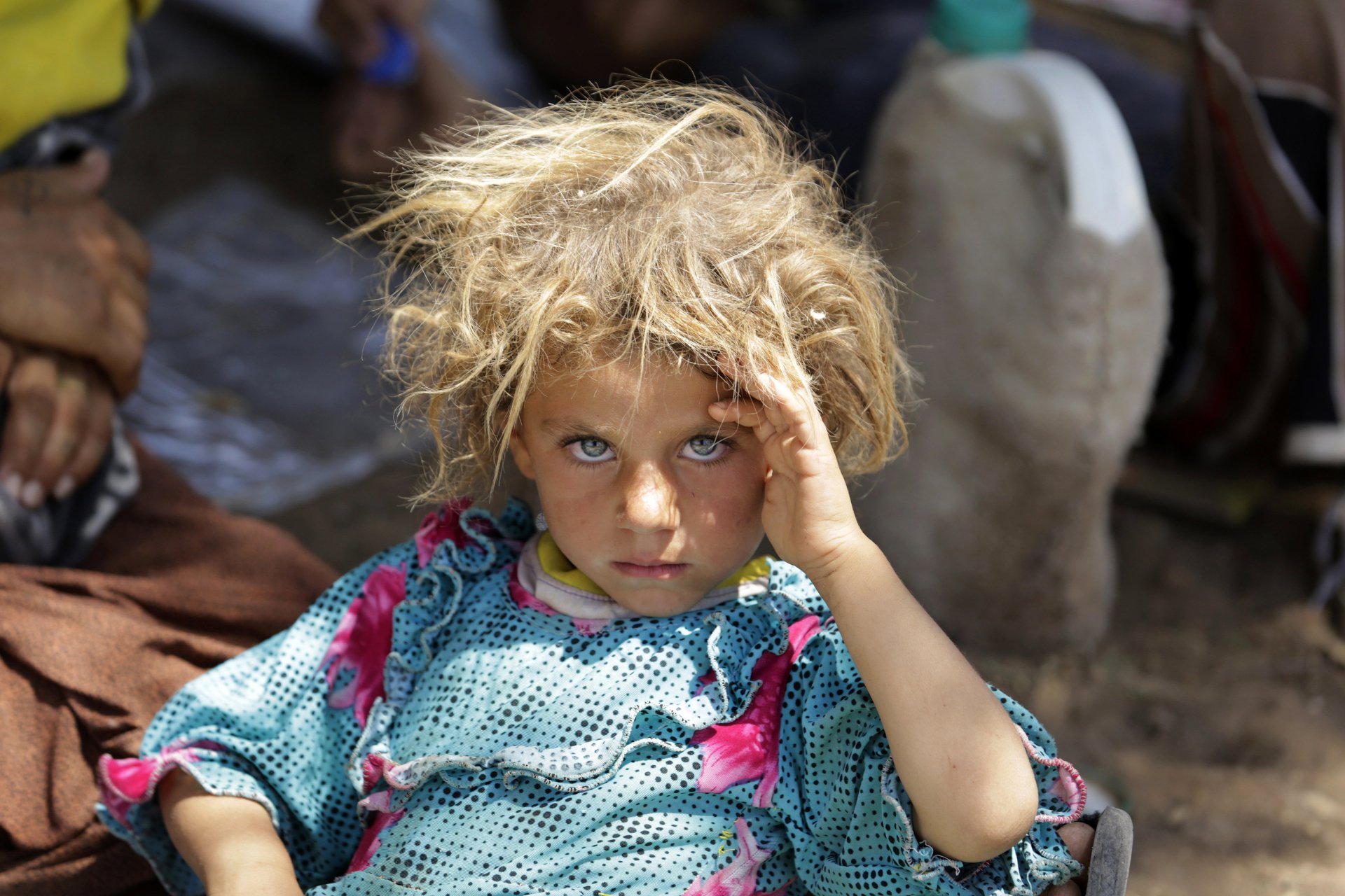 Photography — child with tangled blond hair and bright blue eyes in a patterned dress, sitting on the ground and looking at the camera with one hand to their forehead.