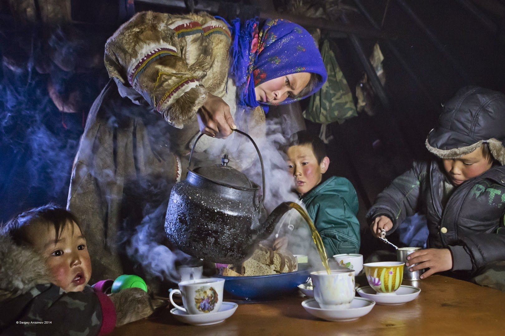 A photography capturing a child and two others gathered around a table as a woman pours hot water from a kettle into cups, creating steam in a cozy indoor setting.