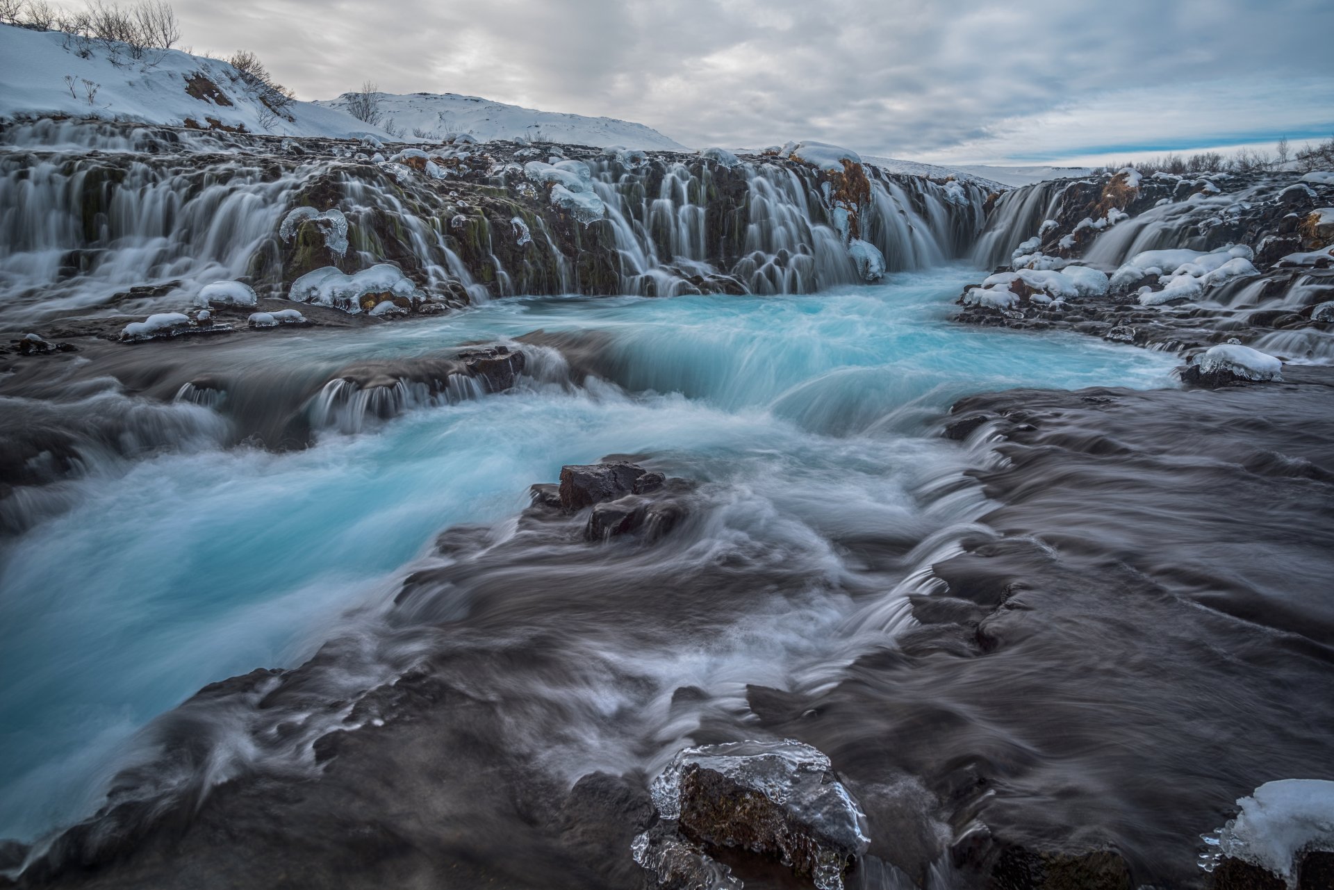 Icelandic waterfall surrounded by icy rocks and winter landscape under a cloudy sky, showcasing nature’s raw beauty.
