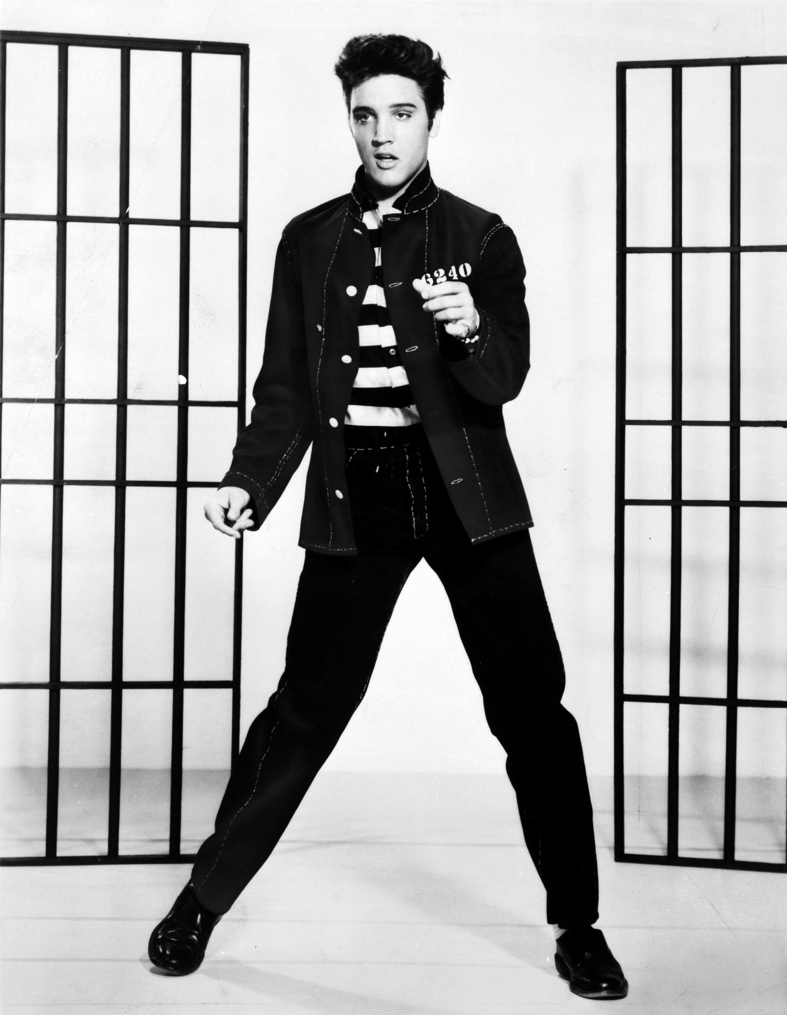 Black-and-white vintage photo of a male American singer in a striped jail costume from the movie Jailhouse Rock, standing between cell bars mid-performance.