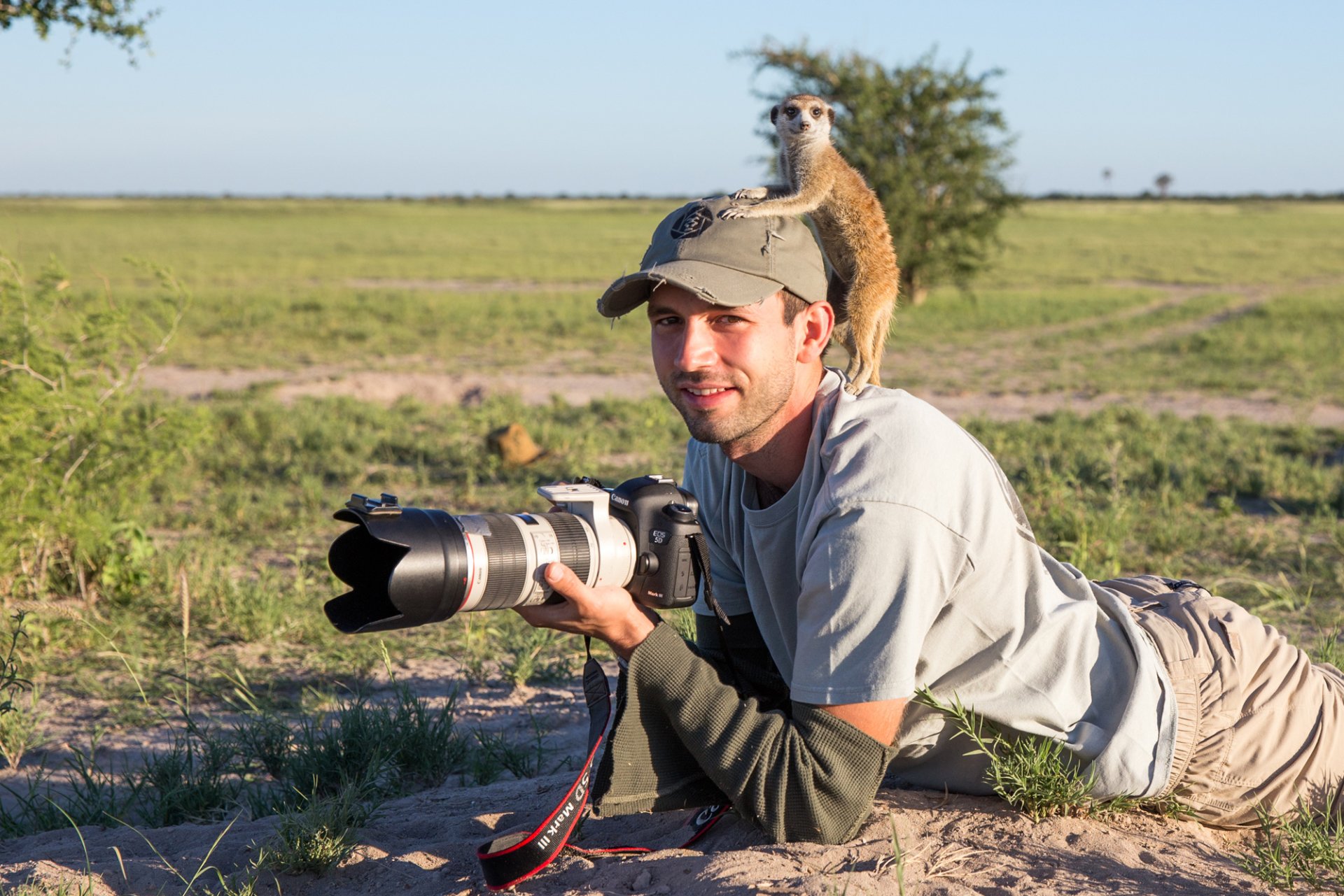 A man lies on the ground holding a camera while a meerkat perches on his shoulder in a grassy open field.