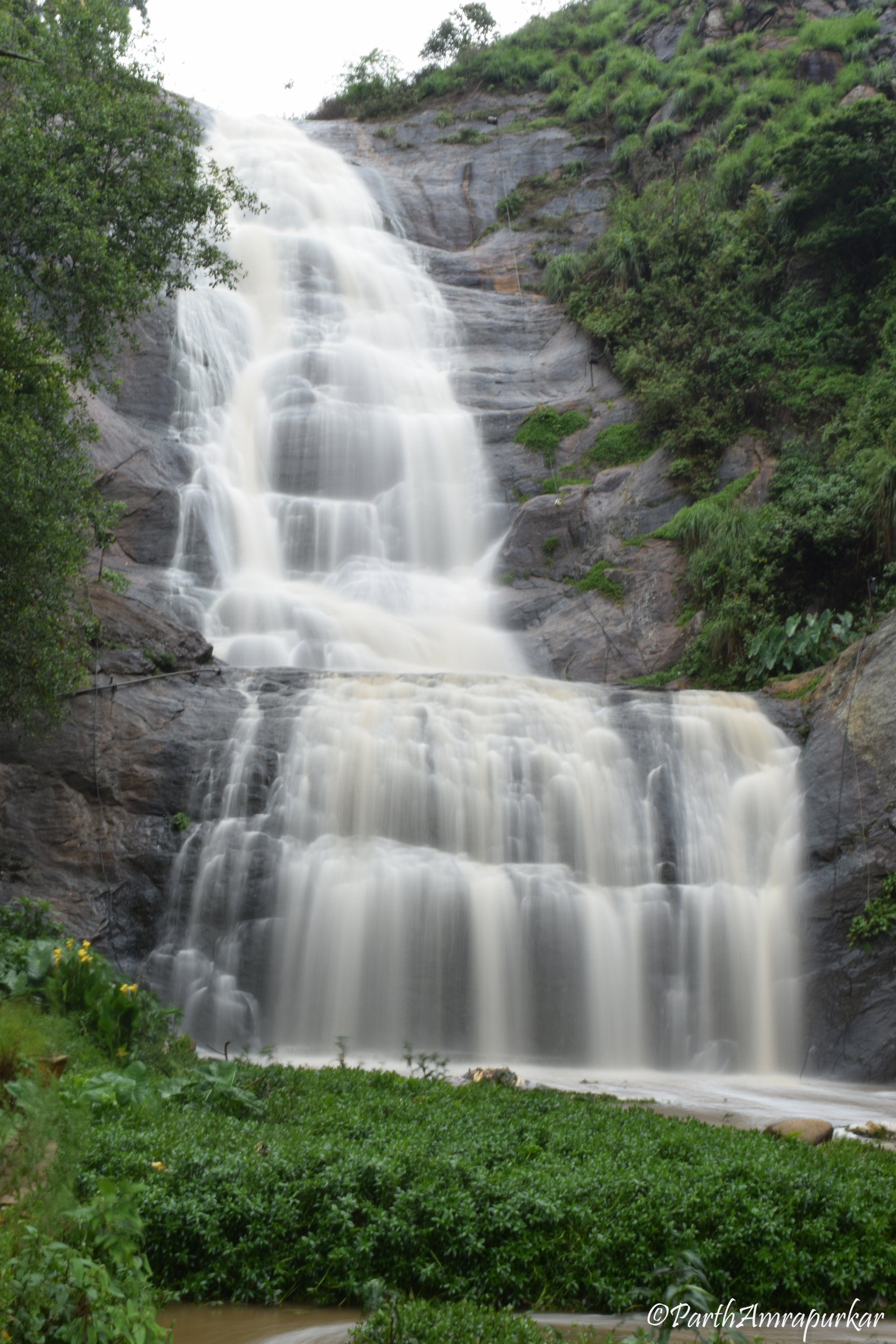 A multi-tiered waterfall cascades down rocky cliffs surrounded by lush green vegetation in a serene natural landscape.