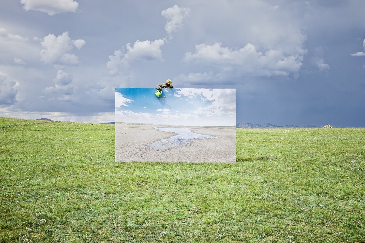 A unique landscape photo features a large canvas displaying a serene beach scene, positioned in an open green field under dramatic clouds.