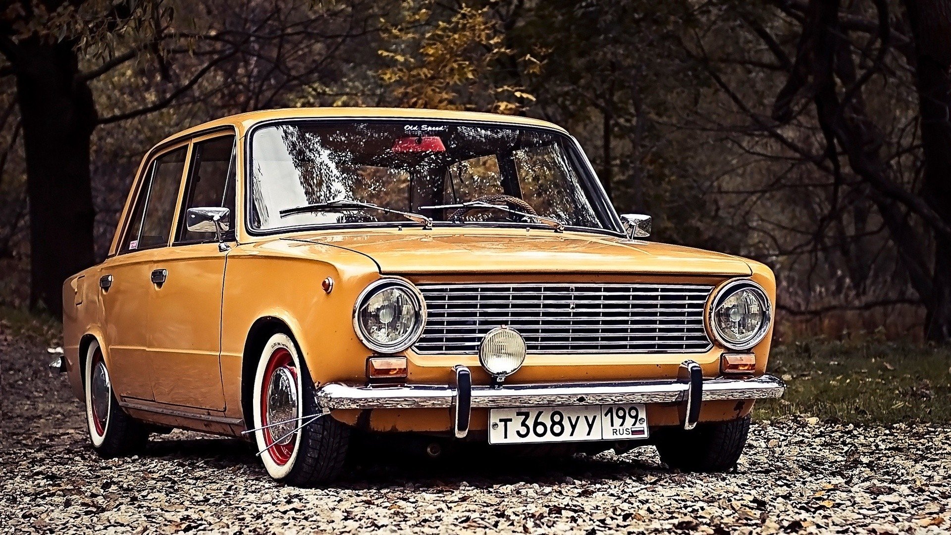 A vintage VAZ-2101, featuring a bright yellow exterior and classic design, is parked on a gravel path surrounded by trees in a scenic setting.