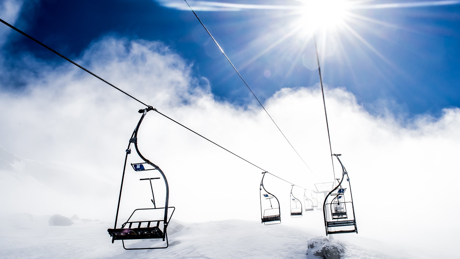 Cable car vehicle chairs suspended above a snowy slope, stretching into bright sun and a clear blue sky.